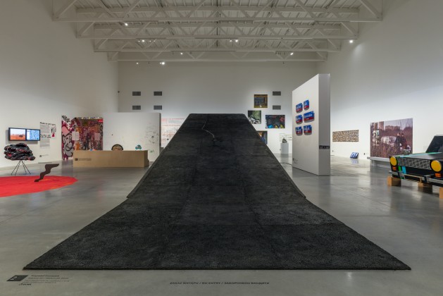 Pas czarnego asfaltu unoszący się do góry przecina niemal całą przestrzeń wystawienniczą Muzeum Sztuki Nowoczesnej, dominując ją. Na około są rozstawione inne prace. 