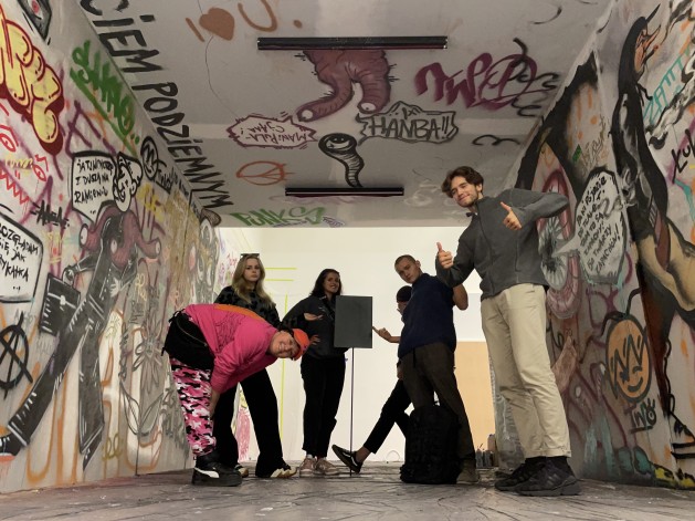 W odtworzonym w Muzeum Sztuki Nowoczesnej podziemnym przejściu wypełnionym kolorowym grafiti stoi sześć, młodych osób pozujących do zdjęcia.