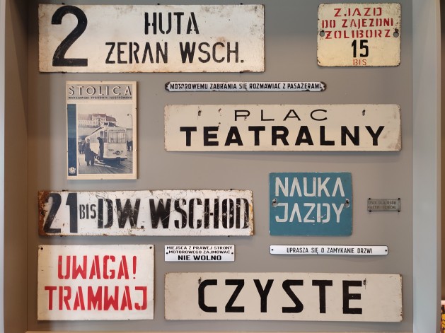 Na ścianie wiszą zabytkowe znaki, plakietki, komunikaty i zdjęcie związane z warszawskim transportem miejskim. Widnieją na nich takie napisy jak: "Czyste", "Plac Teatralny", "Uprasza się o zamykanie drzwi" czy "Uwaga Tramwaj".