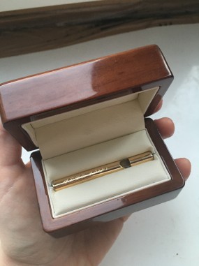 W drewnianym pudełku wypełnionym białym materiałem znajduje się mała, złota piszczałka.