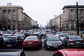 Samochody stoją na parkingu na Placu Konstytucji w Warszawie. Zajmują one większą część placu i dominują krajobraz. Zza samochodów widać rozciągającą się ulicę Marszałkowską.
