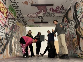 W odtworzonym w Muzeum Sztuki Nowoczesnej podziemnym przejściu wypełnionym kolorowym grafiti stoi sześć, młodych osób pozujących do zdjęcia.