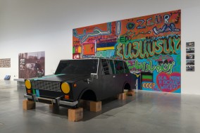 Rzeźba schematycznie, rysunkowo przedstawia czarny samochód naturalnej wielkości, który zamiast kół stoi na pustakach. W tle znajduje się ściana wypełniona kolorowym grafiti.